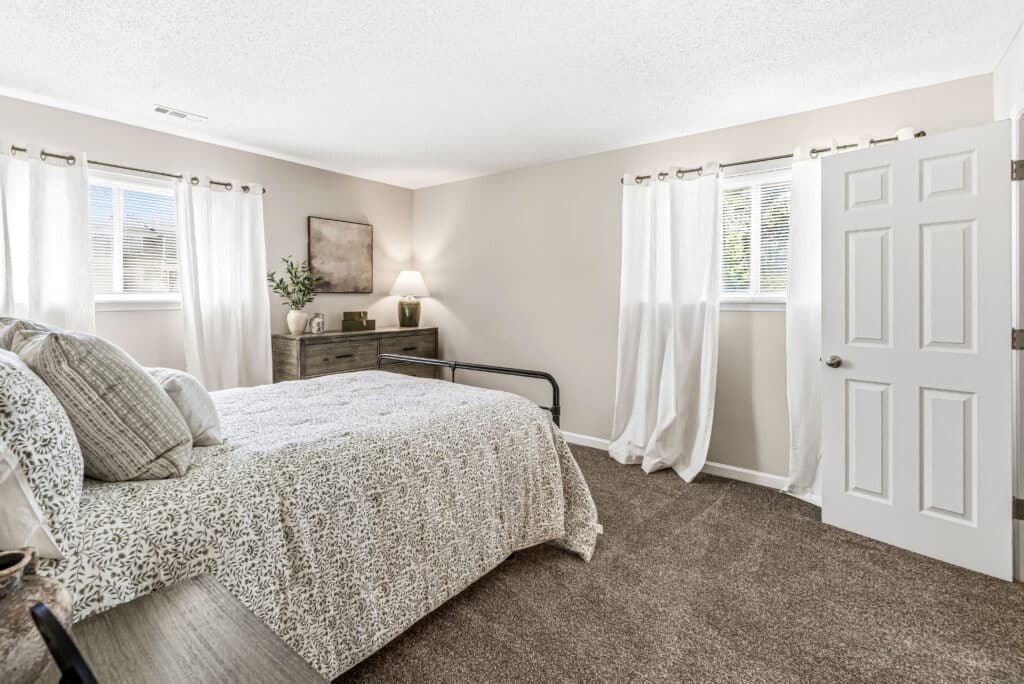 A bedroom with a neatly made bed, a nightstand, a dresser with a lamp and plant, carpeted floor, white walls, and three windows with white curtains.